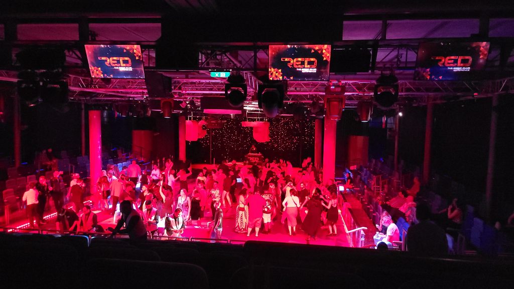Looking down into the high-energy RED Nightclub experience inside Studio B on a Royal Caribbean mega-ship, featuring an active dance floor filled with passengers, glowing red club lighting, a DJ stage, and overhead digital screens displaying the RED nightclub logo.