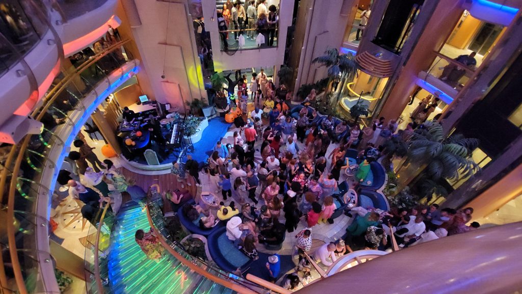 A high-angle aerial view looking straight down into a packed, high-energy evening dance party in the multi-story central atrium of the Radiance of the Seas, featuring a glowing neon staircase and a large crowd dancing.