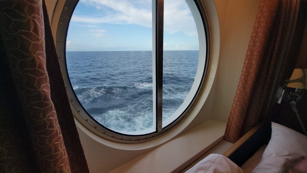 A view from inside a cruise ship stateroom looking out through a large, circular ocean-view porthole window at deep blue sea waves under a partly cloudy sky.