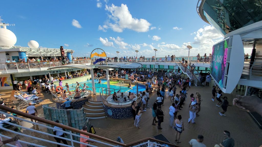A lively daytime sail-away party on the crowded upper pool deck of the Radiance of the Seas, filled with passengers dancing and swimming next to lounge chairs under a blue sky.