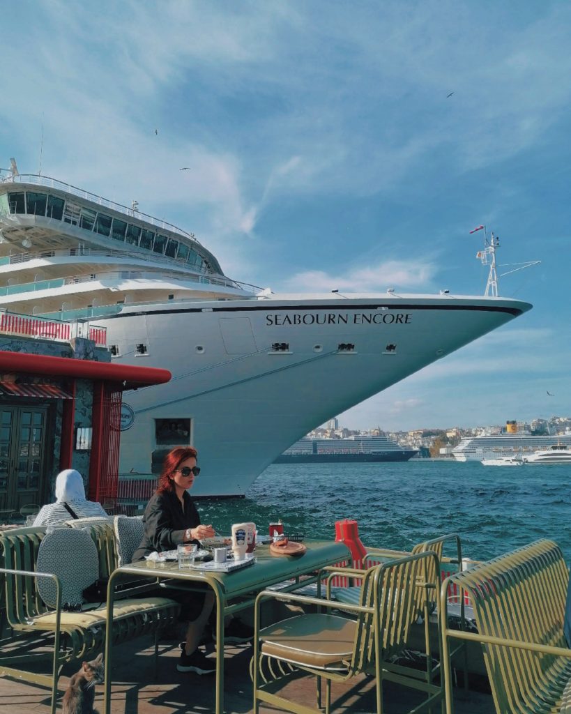 A chic woman dining at a waterfront outdoor café table with the massive white bow of the ultra-luxury cruise ship Seabourn Encore docked directly behind her in Istanbul, Turkey.