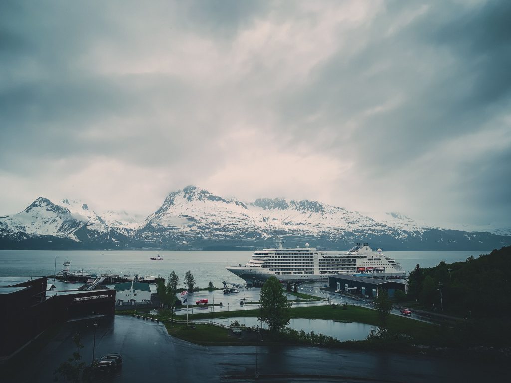 The ultra-luxury cruise ship Silver Muse docked at a northern port surrounded by massive, dramatic snow-capped mountains under an overcast sky.
