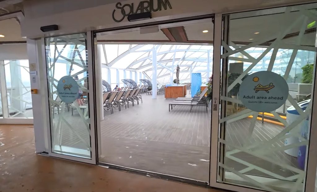Looking through the open sliding glass doors into the modern, sunlit Solarium on Royal Caribbean's Symphony of the Seas, with signs on the glass clearly labeled "SOLARIUM" and "Adult area ahead."