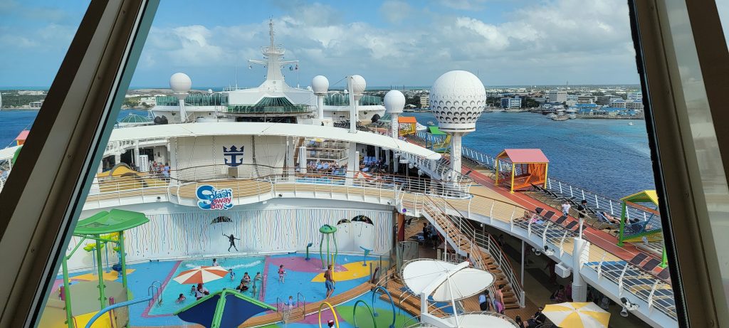 Looking down at the colorful Splashaway Bay children's water park on the sunny top deck of a Royal Caribbean cruise ship, featuring mini water slides, dump buckets, and splashing fountains.
