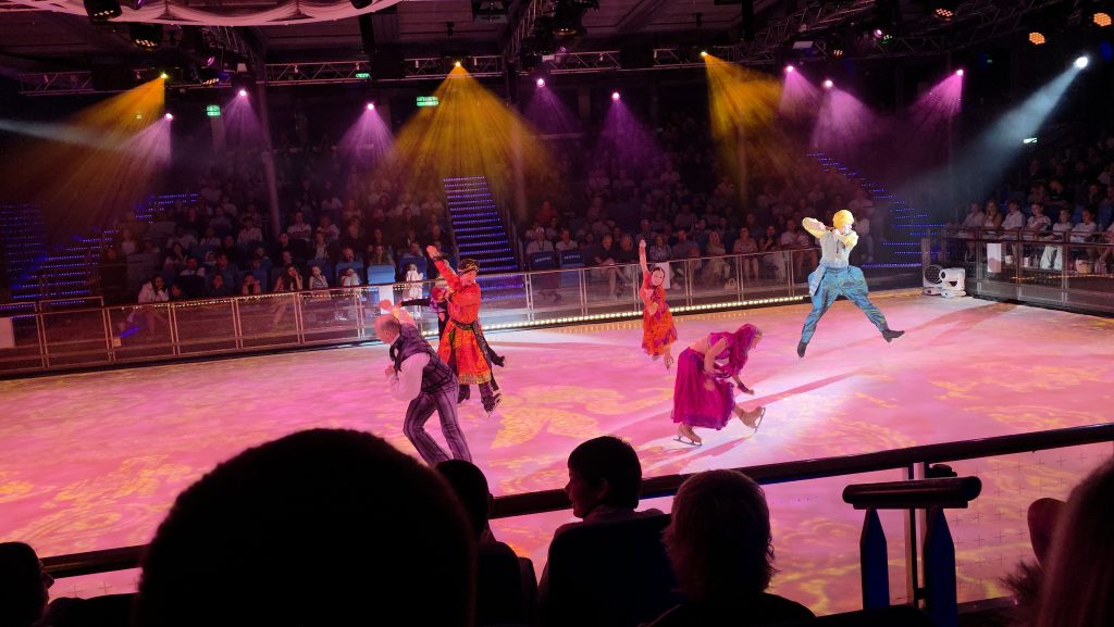 Looking down at a high-energy ice skating performance inside Studio B on a Royal Caribbean cruise ship, featuring costumed professional skaters on a vibrant pink-lit ice rink with one skater frozen mid-air during a dramatic jump under theatrical spotlights.