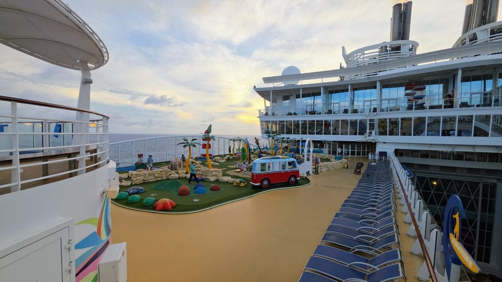 A sunset view of the colorful Symphony Dune mini-golf course on the top deck of a Royal Caribbean mega-ship, featuring whimsical lighthouse and camper-van obstacles with the ocean horizon in the background.
