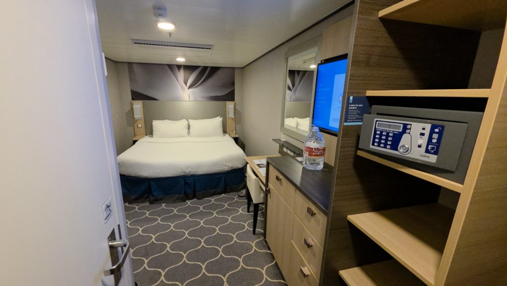 A bright view looking into a modern windowless interior cruise stateroom featuring a neatly made bed, vanity desk, wall-mounted television, and a shelving unit with a digital room safe in the foreground.