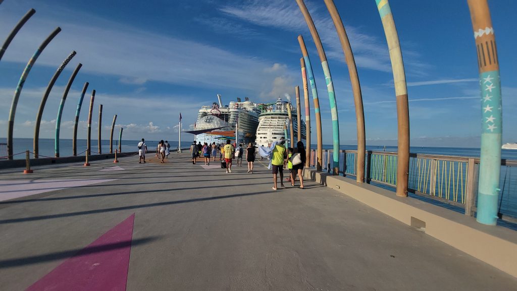 A wide view looking down the arrival pier at Perfect Day at CocoCay, showcasing the massive scale of Royal Caribbean's Oasis-class Utopia of the Seas docked next to the mid-sized Freedom-class Liberty of the Seas under a bright blue sky.