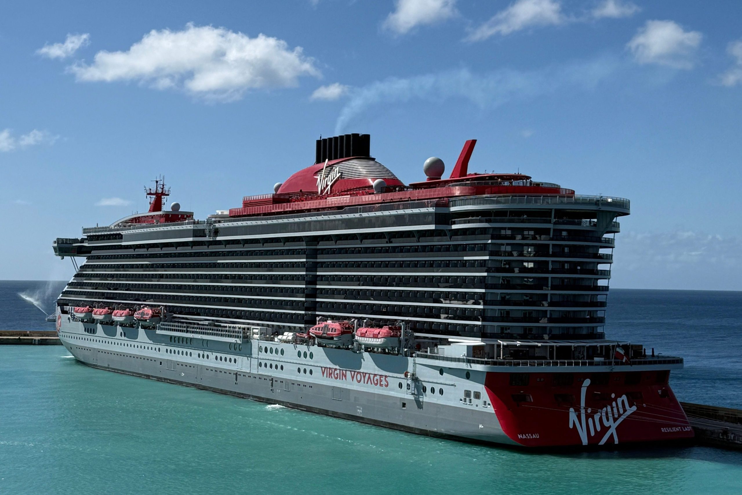 A bright, sunny side-profile view of the Virgin Voyages Resilient Lady cruise ship docked in a vibrant turquoise harbor, highlighting its signature red funnel and sleek grey hull design.